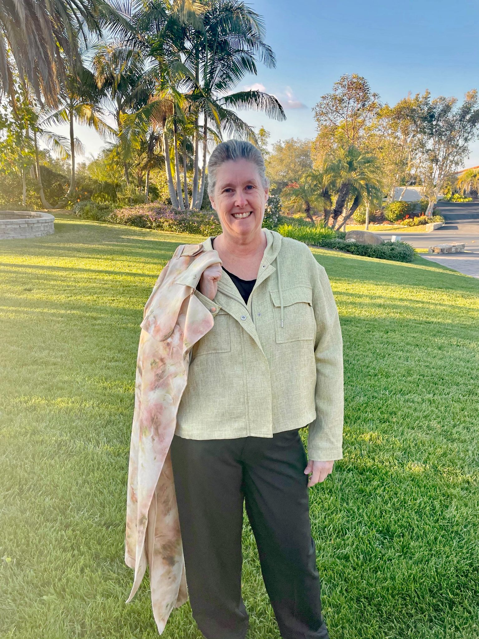 Person standing on grass, wearing a light jacket and holding a scarf. Trees and a road are visible in the background under a clear sky.
